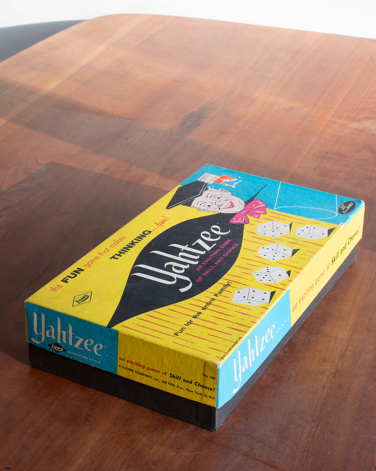 A boxed copy of the Yahtzee game sits on a wooden tabletop. The box is yellow and turquoise with black and white illustrations and the title 'Yahtzee' prominently visible.