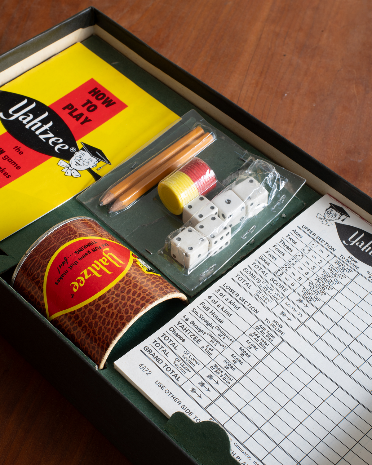 A Yahtzee game set displayed inside its dark green box on a wooden table. The set includes a yellow 'How to Play' booklet, a brown crocodile-print paper cup, wooden dice, a plastic bag with two wooden sticks and white dice, and a printed score sheet.