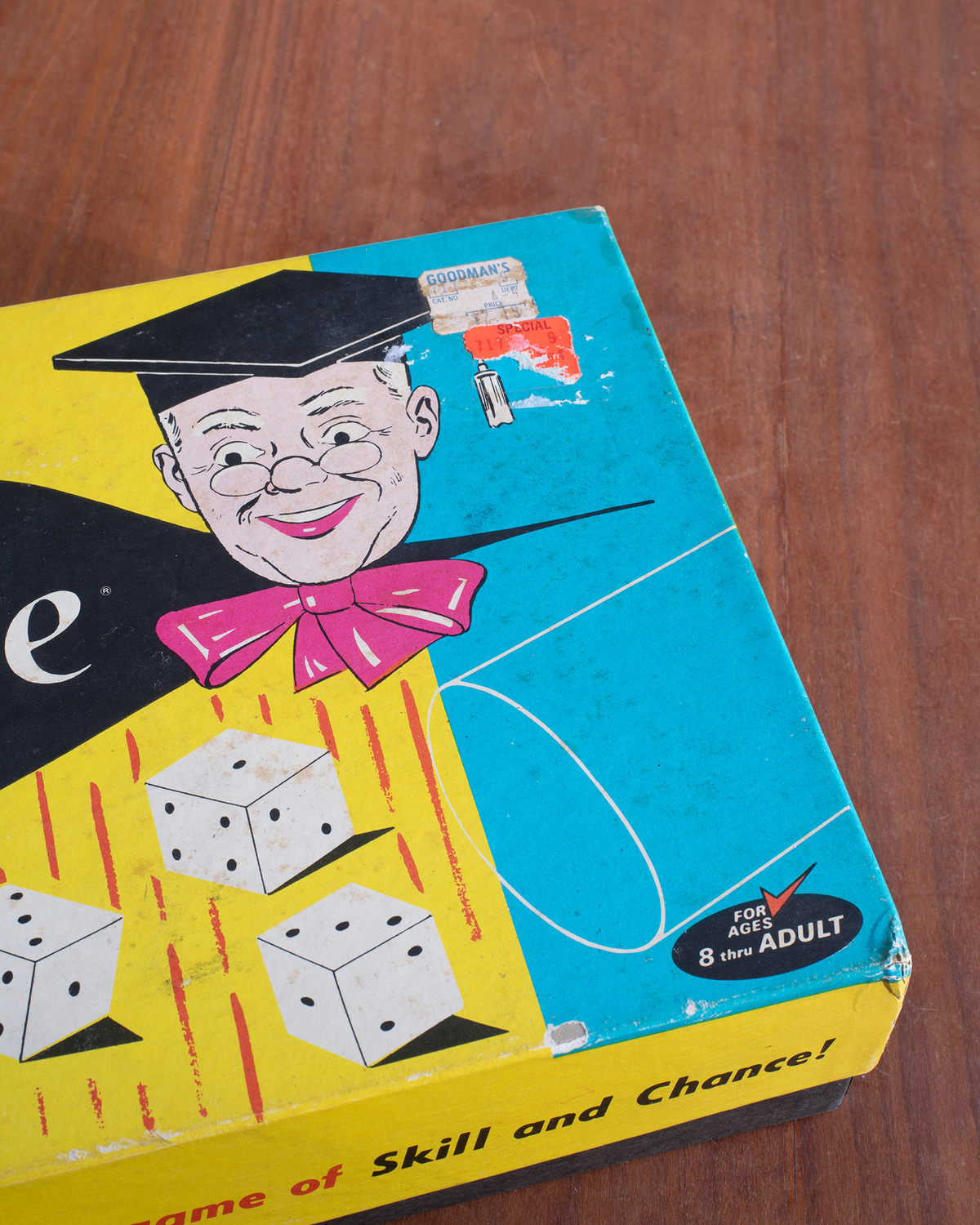 Vintage board game box titled 'Game of Skill and Chance!' shown on a wooden table. The box features a bright yellow and blue design with an illustration of a graduate wearing a black cap and pink bow tie, along with white dice.