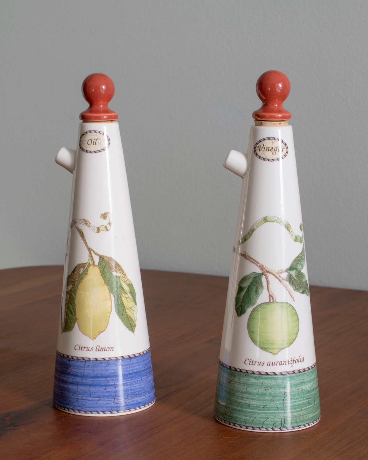 Two decorative ceramic bottles with conical shapes, featuring citrus fruit illustrations and blue and green textured bands at the base, displayed on a wooden surface against a gray wall.