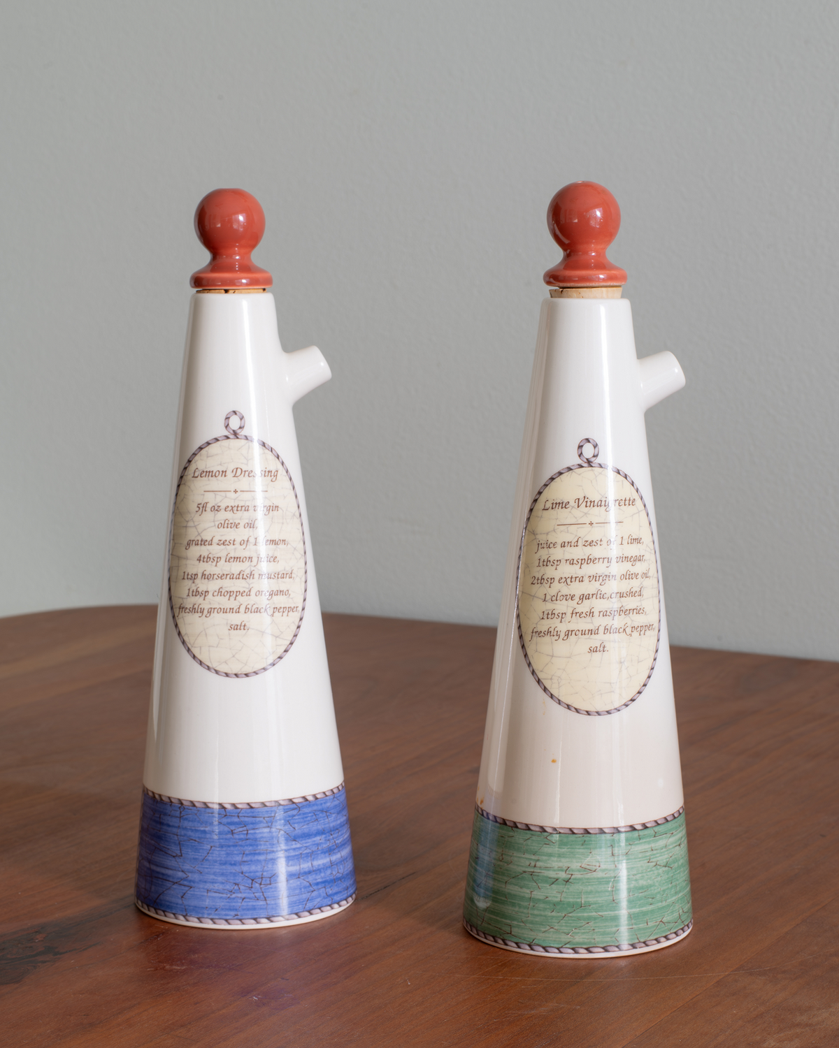 Two decorative ceramic oil bottles with white bodies, red wooden stoppers, and painted bases, displayed on a wooden surface against a gray wall.