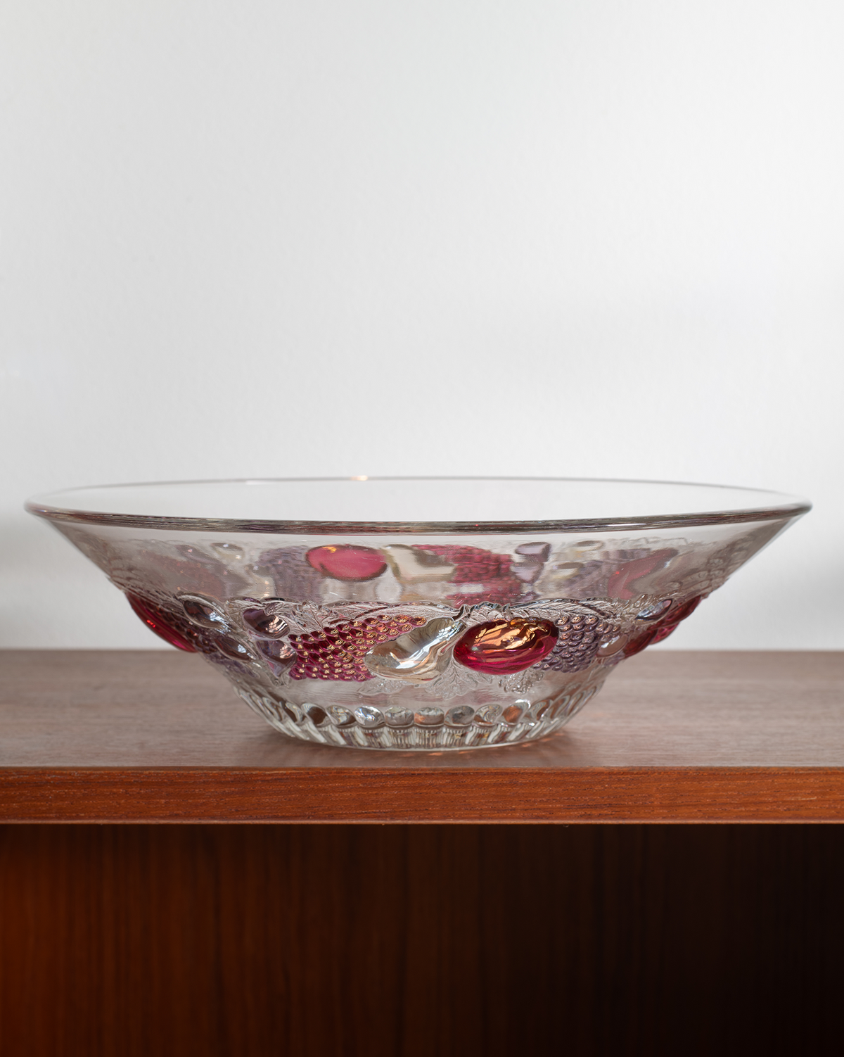 A clear glass bowl with embossed fish patterns and red accents, resting on a wooden shelf against a plain light background.