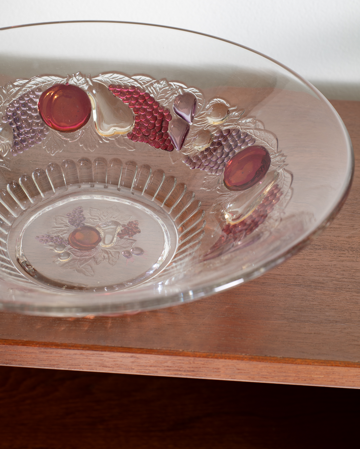 A clear glass serving bowl with embossed floral and fruit motifs in muted red, purple, and beige tones, resting on a wooden surface.