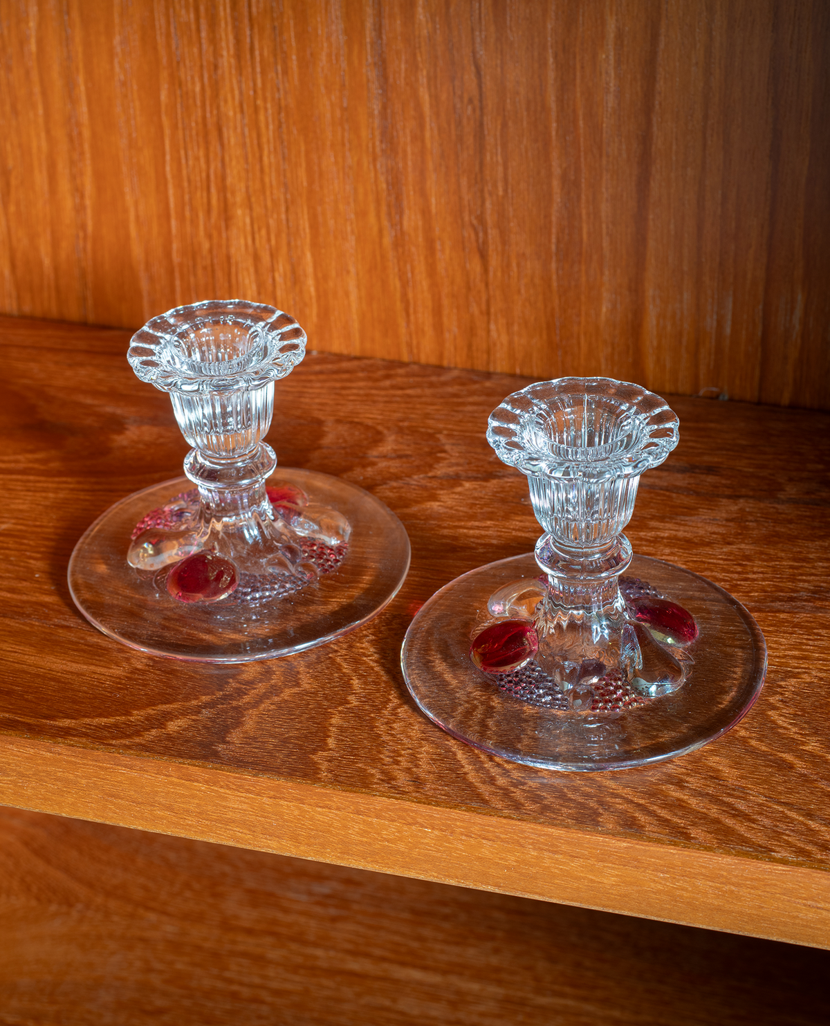 Two clear glass candlestick holders with red and gold decorative accents, displayed on a wooden shelf against a wood-paneled background.