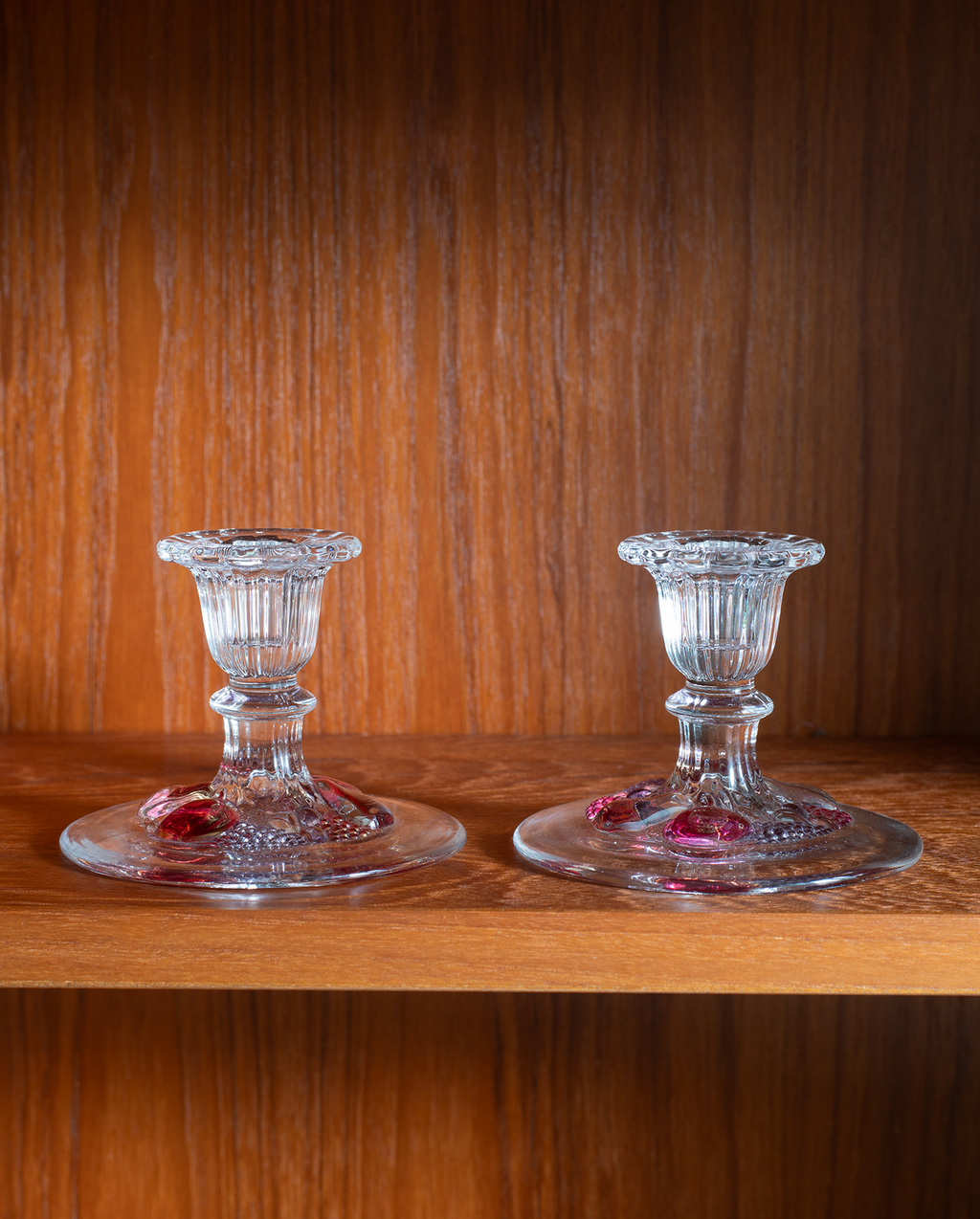 Two clear glass candlestick holders with red floral accents, displayed on a wooden shelf against a wood-paneled background.