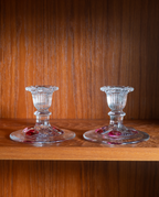 Two clear glass candlestick holders with red floral accents, displayed on a wooden shelf against a wood-paneled background.