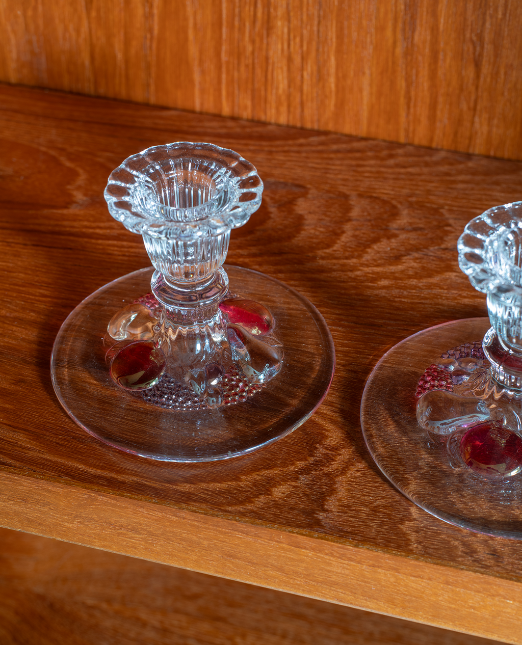 A close-up of a clear glass candle holder with a flared stem and round base, placed on a warm brown wooden surface. Part of a second matching holder is visible at the right edge.