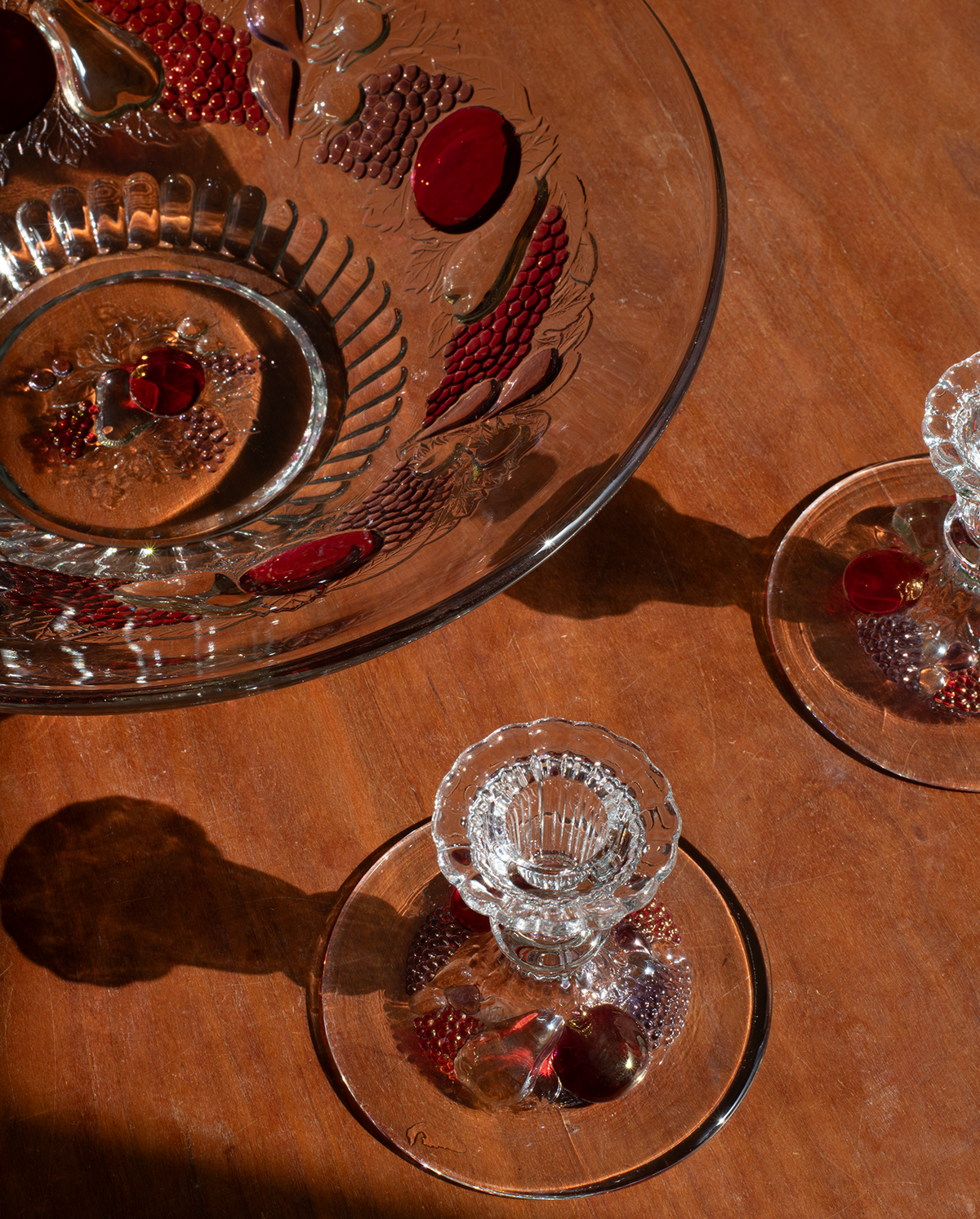 A set of clear glass tableware on a warm brown wooden table, including a large decorative bowl, a small pedestal candle holder, and matching coasters, with warm amber lighting casting strong shadows.