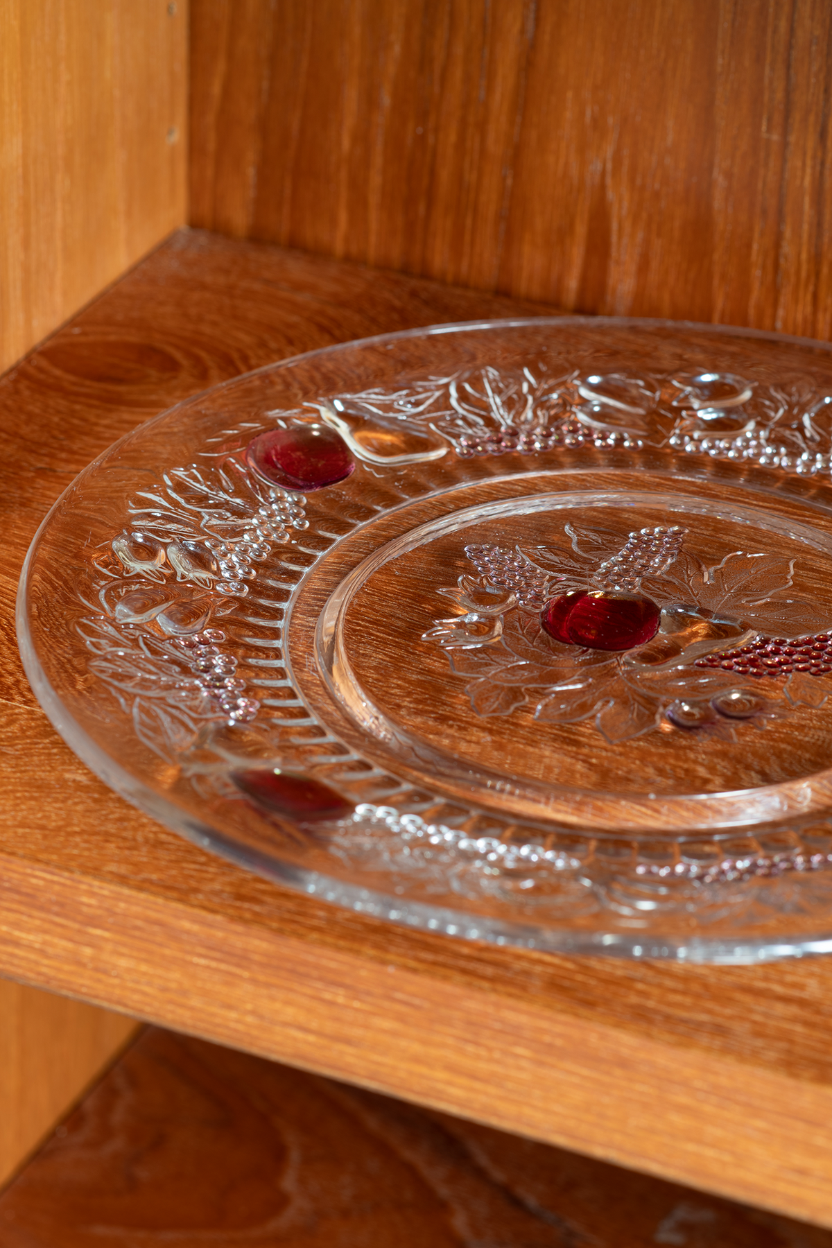 A round clear glass plate with embossed floral and vine patterns, featuring three small red glass accents, displayed on a wooden shelf.