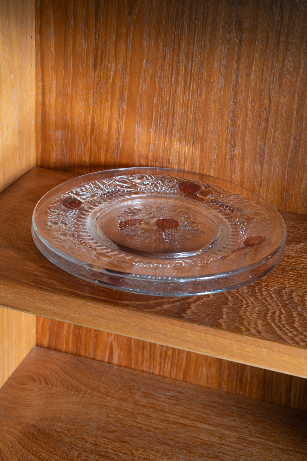 A round clear glass plate with embossed floral patterns and small amber-colored inclusions, displayed on a wooden shelf.