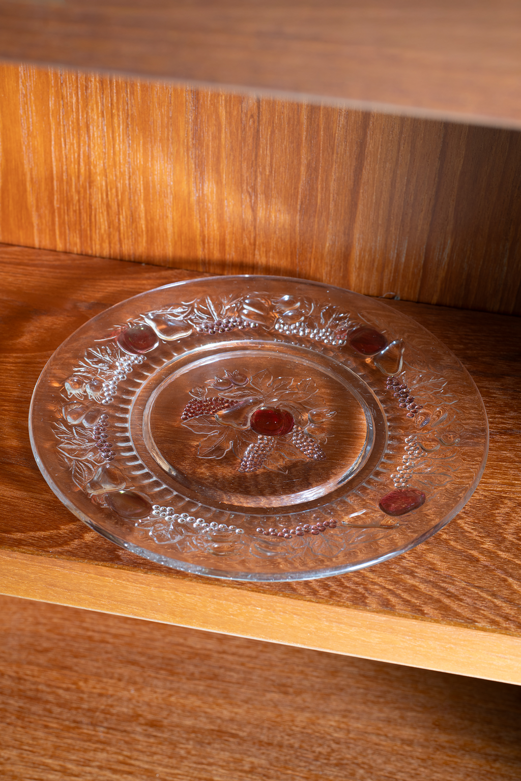 A clear glass plate with embossed floral and grape designs, featuring small red oval inlays, displayed on a wooden shelf.