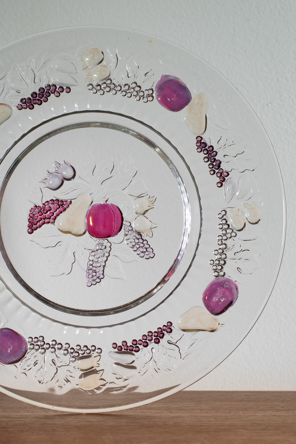 A clear glass decorative plate with a silver rim, featuring embossed floral patterns and scattered purple, pink, and white decorative stones or beads arranged on the surface, photographed against a white wall with a wooden surface below.