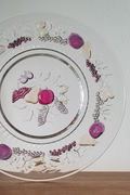 A clear glass decorative plate with a silver rim, featuring embossed floral patterns and scattered purple, pink, and white decorative stones or beads arranged on the surface, photographed against a white wall with a wooden surface below.