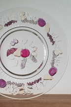 A clear glass decorative plate with a silver rim, featuring embossed floral patterns and scattered purple, pink, and white decorative stones or beads arranged on the surface, photographed against a white wall with a wooden surface below.