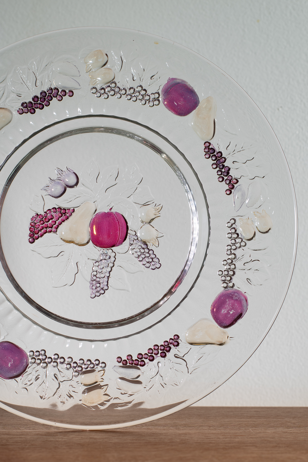 A clear glass decorative plate with a silver rim, featuring embossed floral patterns and scattered purple, pink, and white decorative stones or beads arranged on the surface, photographed against a white wall with a wooden surface below.