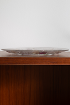 A round clear glass plate with a subtle purple and gold floral pattern, resting on a dark wood shelf against a plain light gray wall.