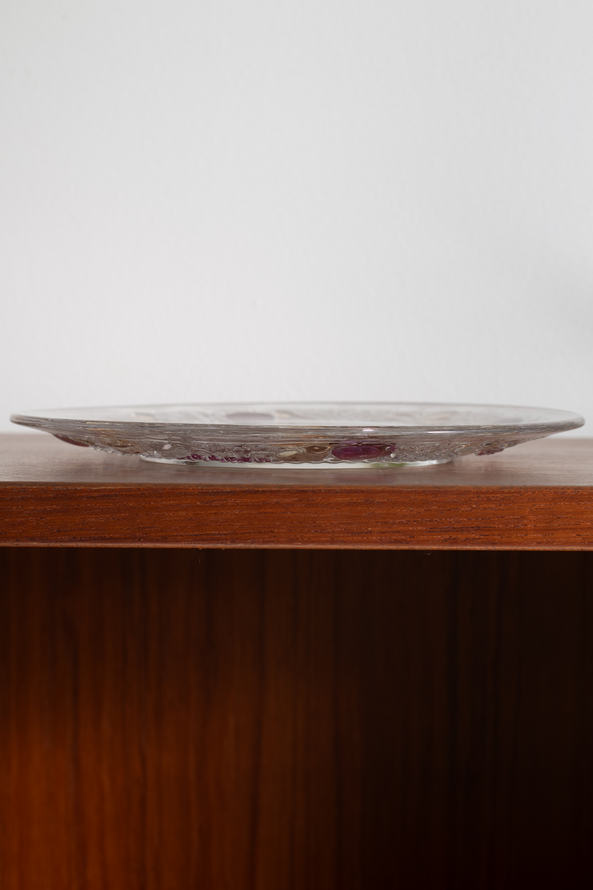 A round clear glass plate with a subtle purple and gold floral pattern, resting on a dark wood shelf against a plain light gray wall.