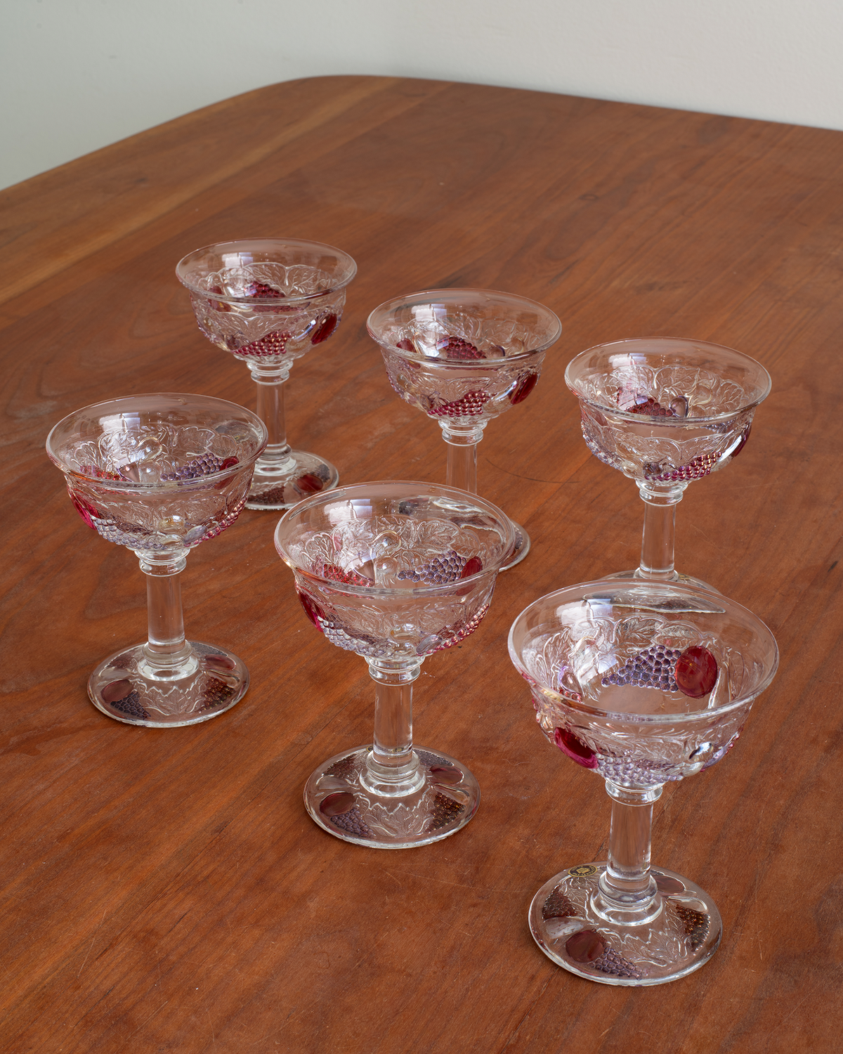 Six clear cut-glass cocktail coupe glasses with red grapevine patterns, arranged on a wooden table.