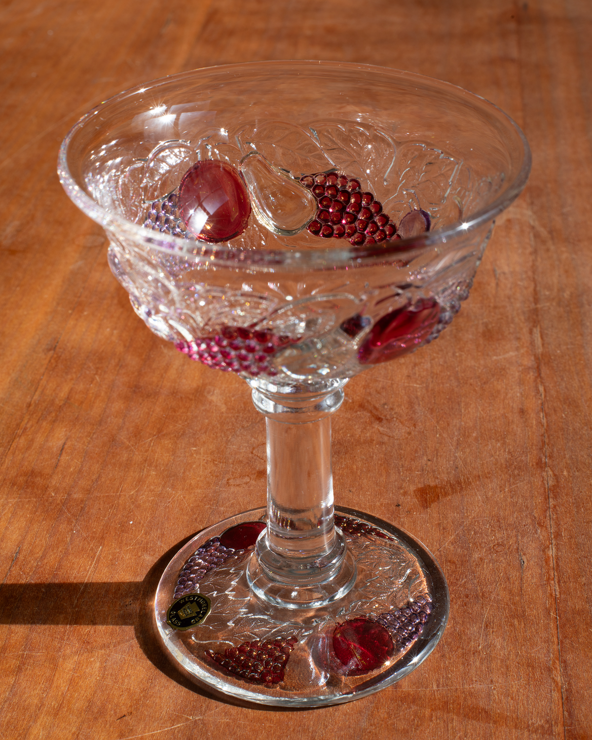 A clear cut-glass coupe-shaped goblet with a stem and circular base, shown on a wooden tabletop with red berries placed inside and reflected on the glass.