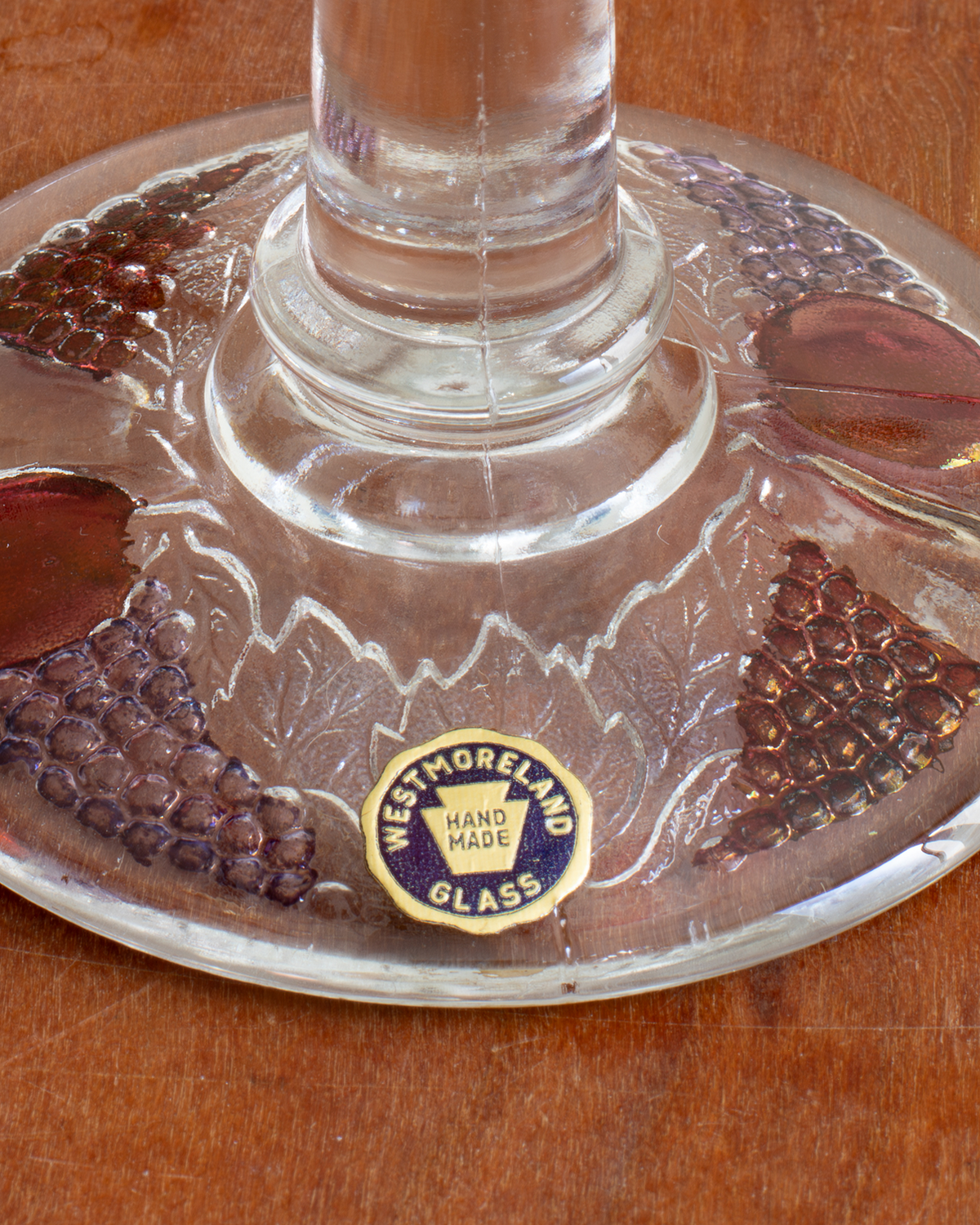 A close-up of a Westmoreland Hand Made clear glass footed candlestick base on a wooden surface, showing the clear glass design, etched details, and a blue and gold circular brand sticker.