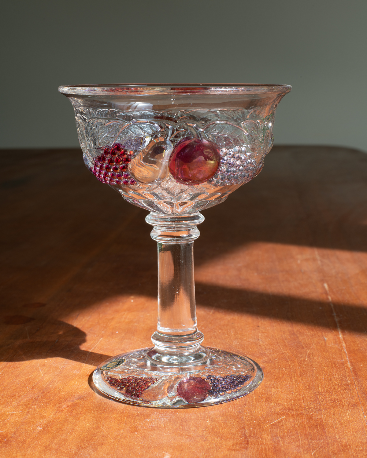 A clear glass goblet with an embossed grapevine pattern and embedded red and purple grapes, set on a wooden table in direct sunlight.