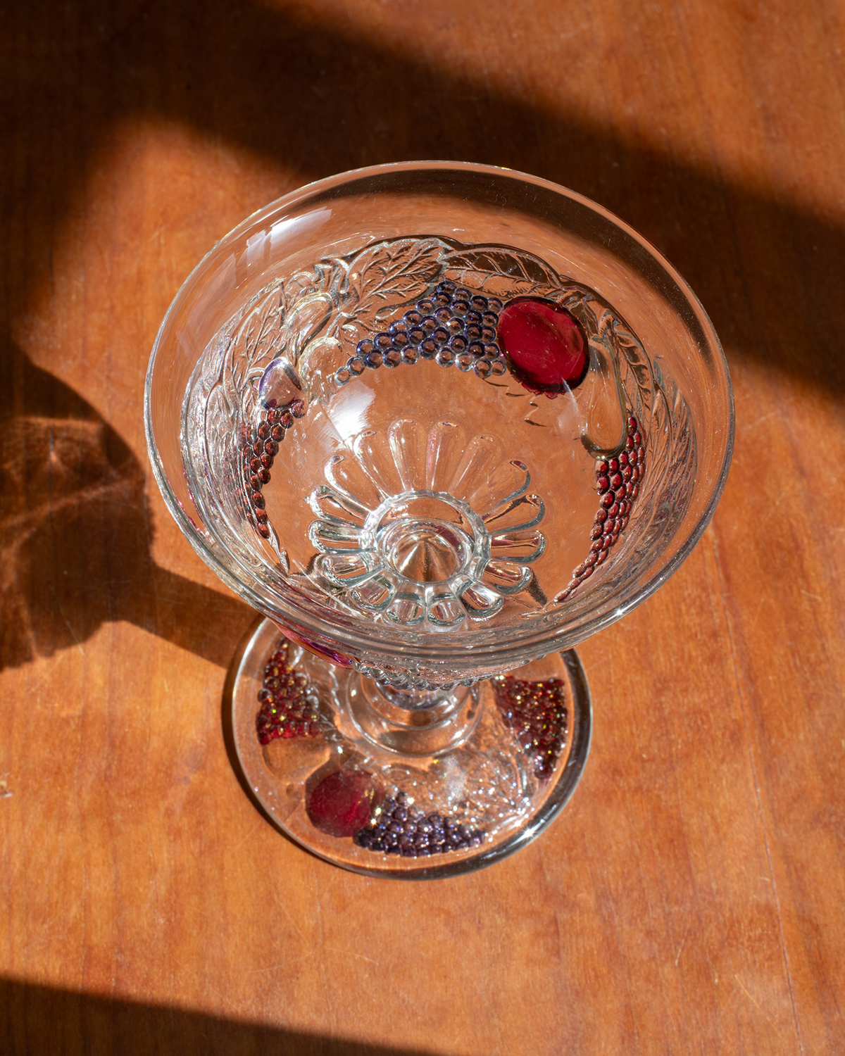 A clear glass dessert goblet with embossed grape and leaf motifs, photographed on a warm wooden surface in strong natural sunlight with deep shadows.