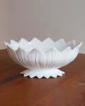 A white ceramic lotus-shaped bowl with a pedestal base, displayed on a wooden tabletop against a neutral beige background.