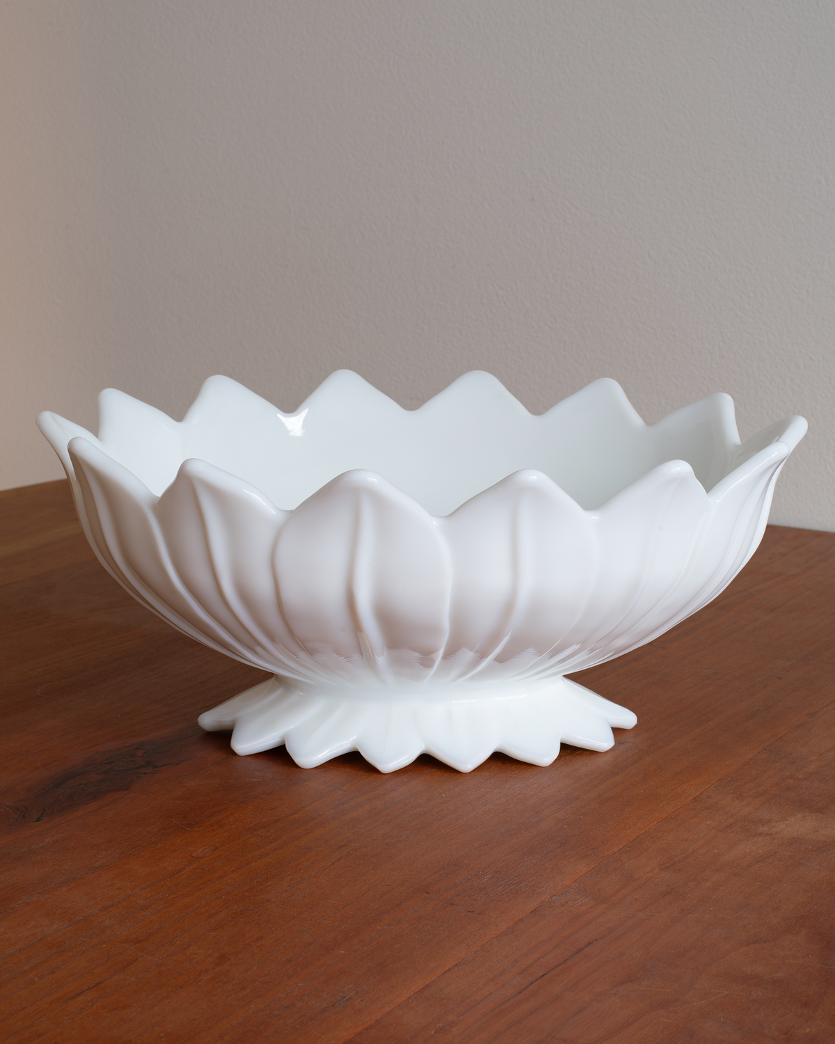 A white ceramic lotus-shaped bowl with a pedestal base, displayed on a wooden tabletop against a neutral beige background.
