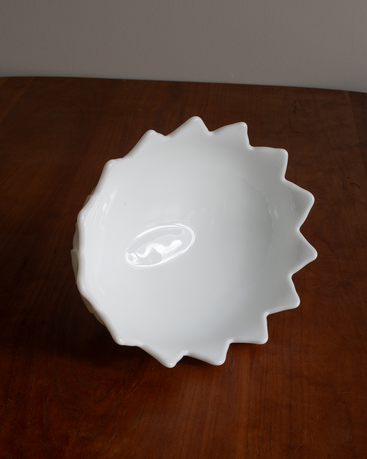 A white ceramic bowl with a distinctive serrated, starburst-like rim, shown on a dark brown wooden tabletop against a neutral gray wall.