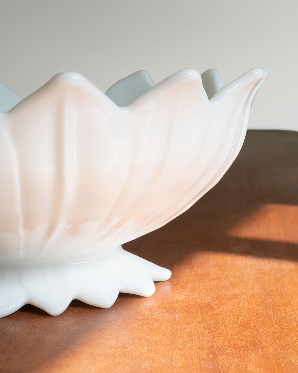 A close-up of a glossy white ceramic bowl with a flared, lotus-like rim, resting on a warm brown wooden surface with soft sunlight and shadows.
