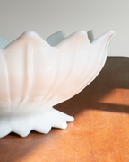 A close-up of a glossy white ceramic bowl with a flared, lotus-like rim, resting on a warm brown wooden surface with soft sunlight and shadows.