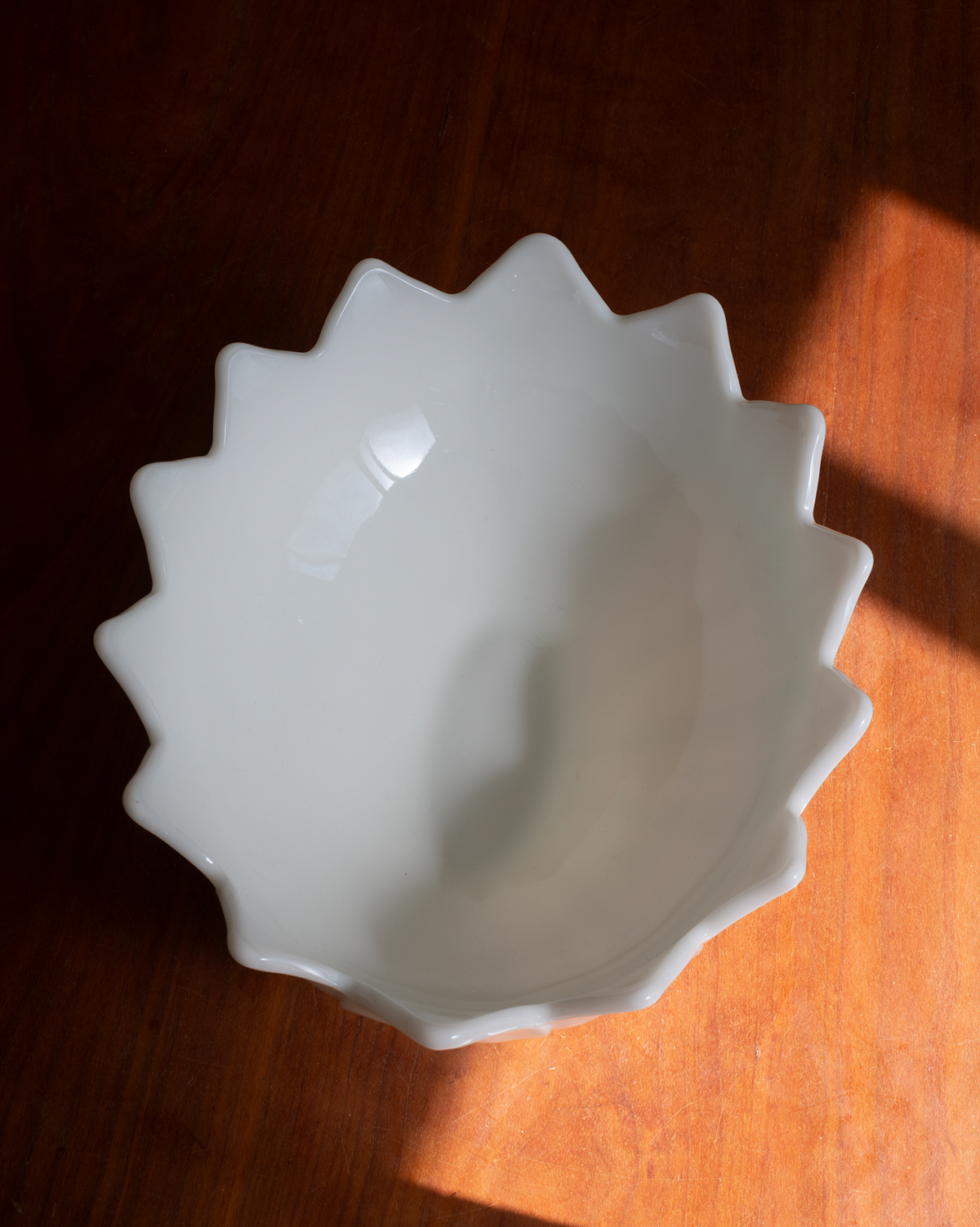 A white ceramic bowl with a scalloped, starburst-like rim, photographed from above on a warm brown wooden surface with sunlight casting shadows.
