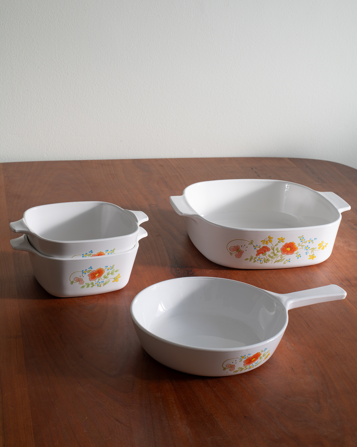 A set of white ceramic cookware with a small orange and yellow floral pattern, displayed on a wooden table against a plain light wall.
