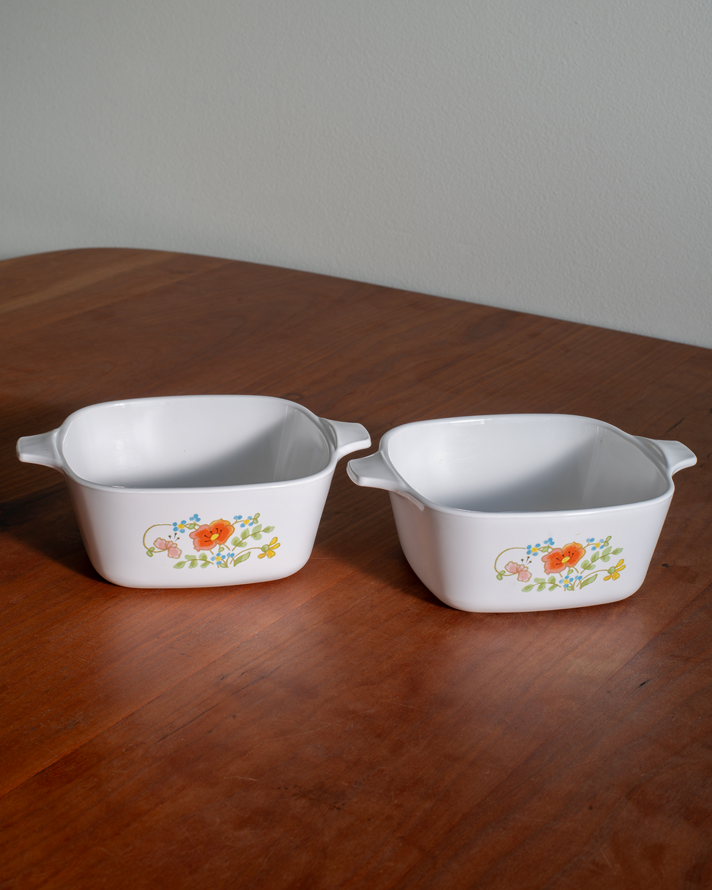 Two white square ceramic baking dishes with side handles, decorated with a small orange and yellow floral pattern, sitting on a wooden table.
