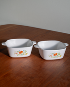 Two white square ceramic baking dishes with side handles, decorated with a small orange and yellow floral pattern, sitting on a wooden table.