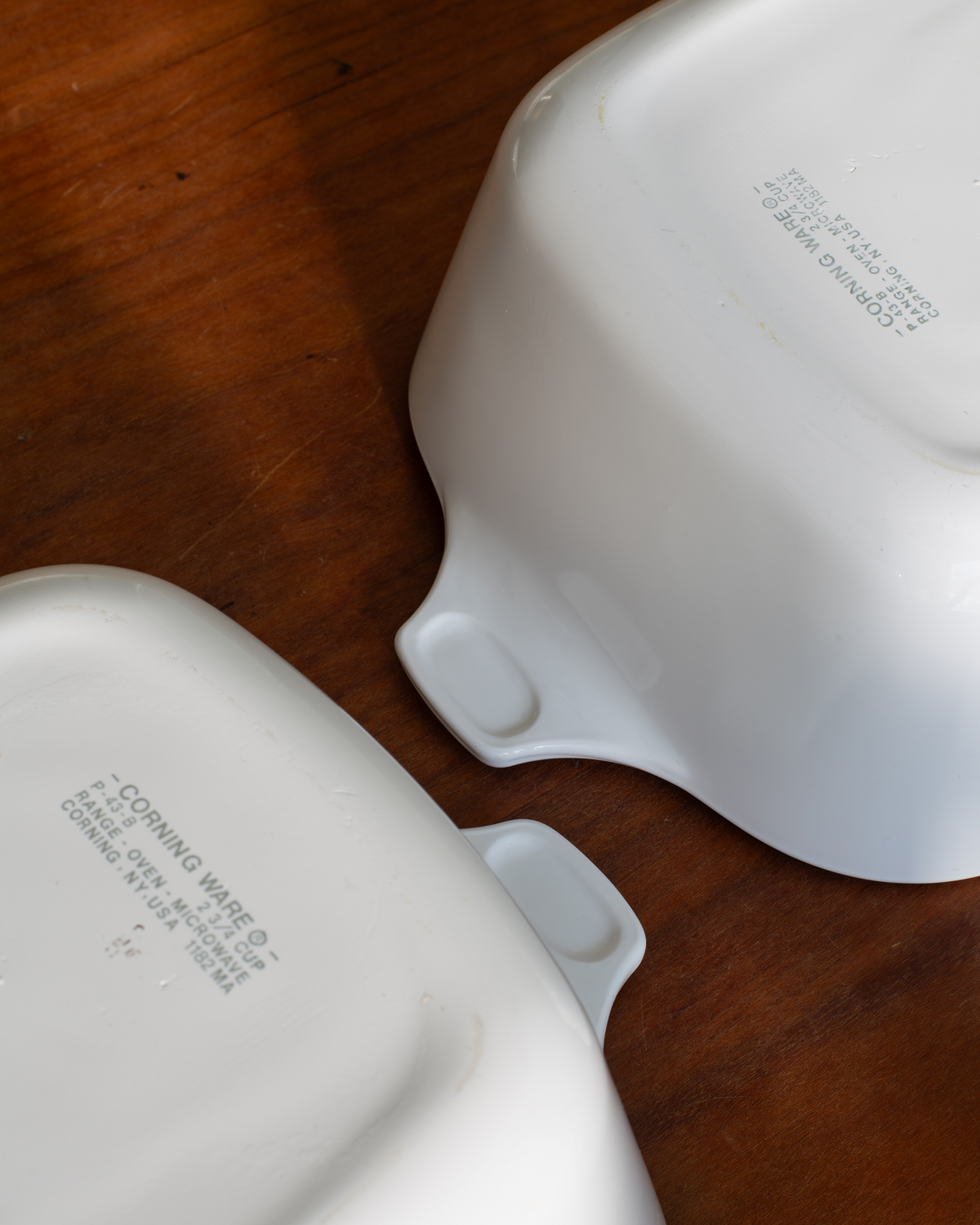 Two white ceramic baking dishes with small side handles, shown upside down on a wooden surface. Faint gray printed text is visible on the inside of the dishes.