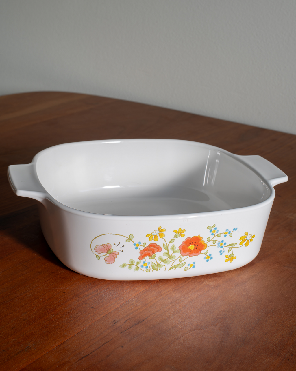 A white rectangular casserole dish with side handles, decorated with a colorful floral pattern of orange, yellow, and pink flowers with green leaves, resting on a wooden table.