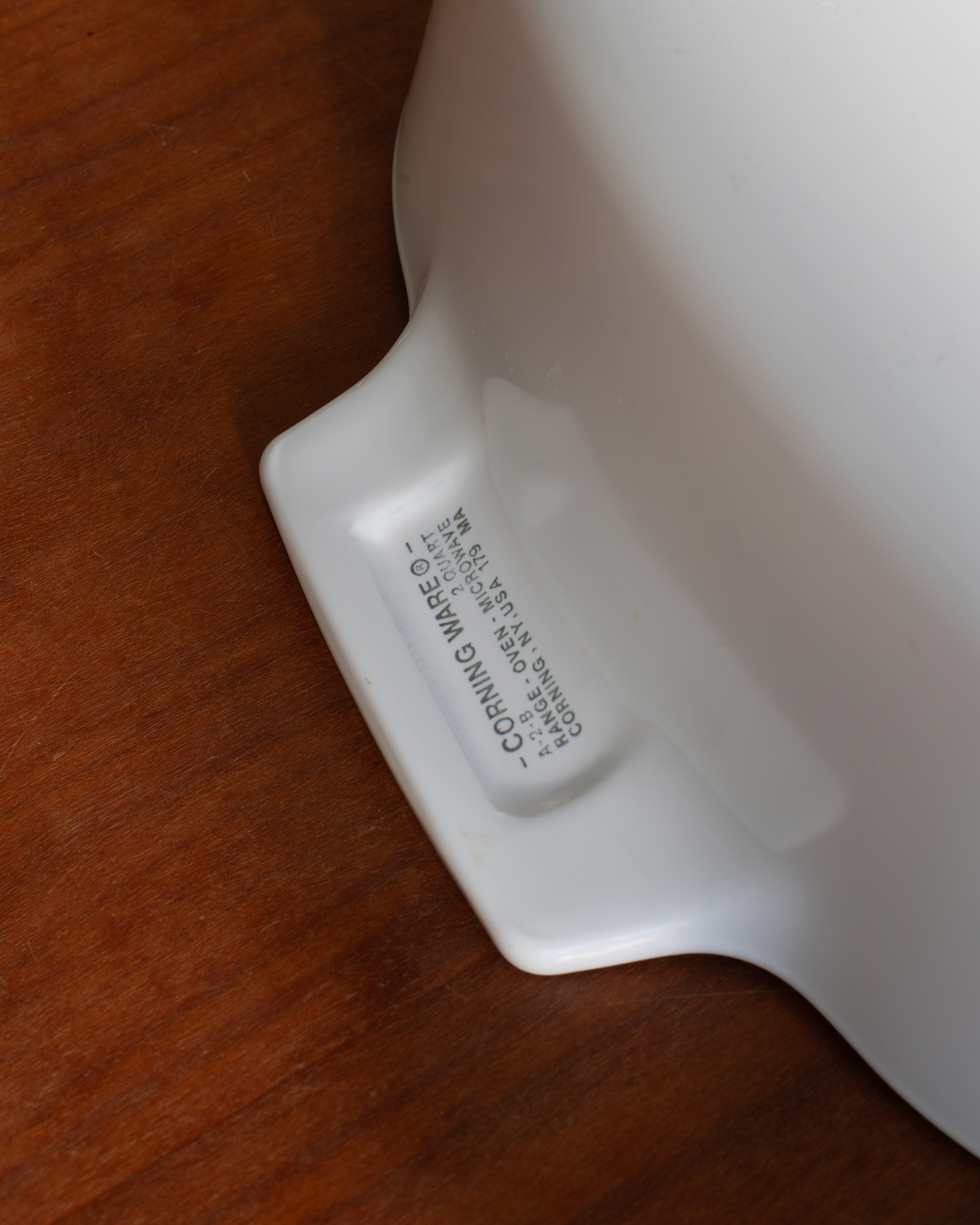 Close-up of the bottom corner of a white ceramic bowl or pitcher resting on a brown wooden surface, showing printed warning text on the base.