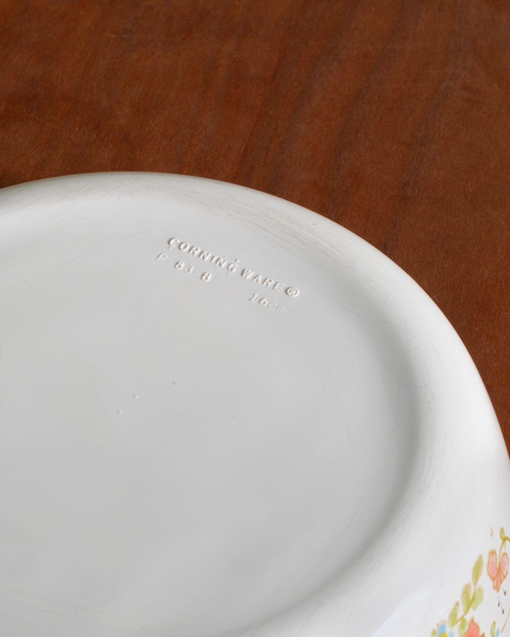Close-up of a white ceramic plate showing the embossed 'Corning Ware' brand mark on the bottom, set against a brown wooden background.