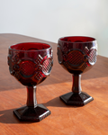 A pair of vintage dark red glass goblets with embossed fish-scale patterns, displayed on a wooden tabletop against a neutral light background.