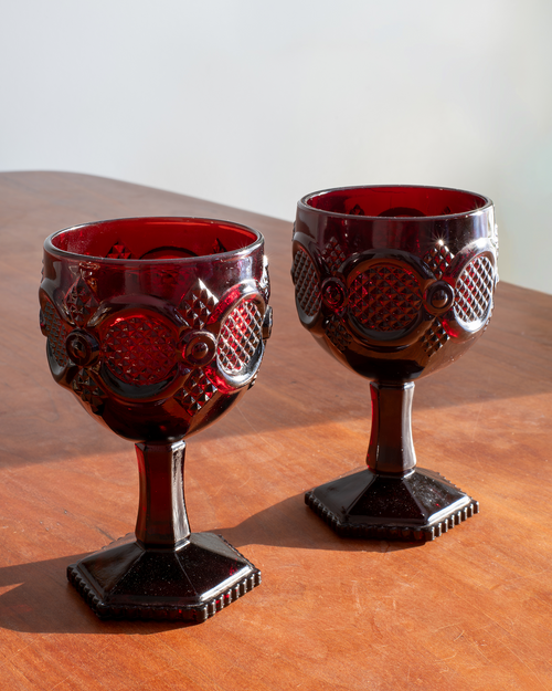 A pair of vintage dark red glass goblets with embossed fish-scale patterns, displayed on a wooden tabletop against a neutral light background.