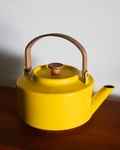 A bright yellow enamel teapot with a brown leather handle and a wooden lid knob, resting on a dark wooden surface against a plain light gray wall.