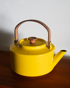 A bright yellow enamel teapot with a brown leather handle and a wooden lid knob, resting on a dark wooden surface against a plain light gray wall.
