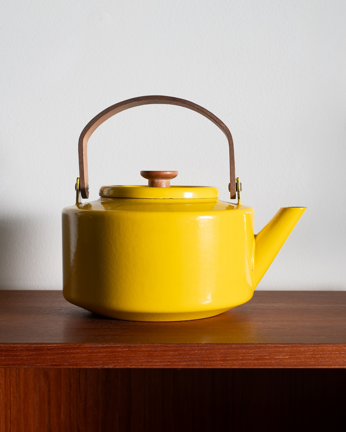 A bright yellow teapot with a brown leather handle and brass valve hardware, sitting on a dark wooden shelf against a plain light gray wall.