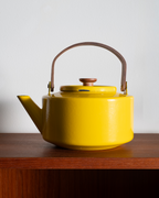 A bright yellow enamel teapot with a brown leather handle and wooden lid knob, sitting on a dark wooden shelf against a plain light gray wall.