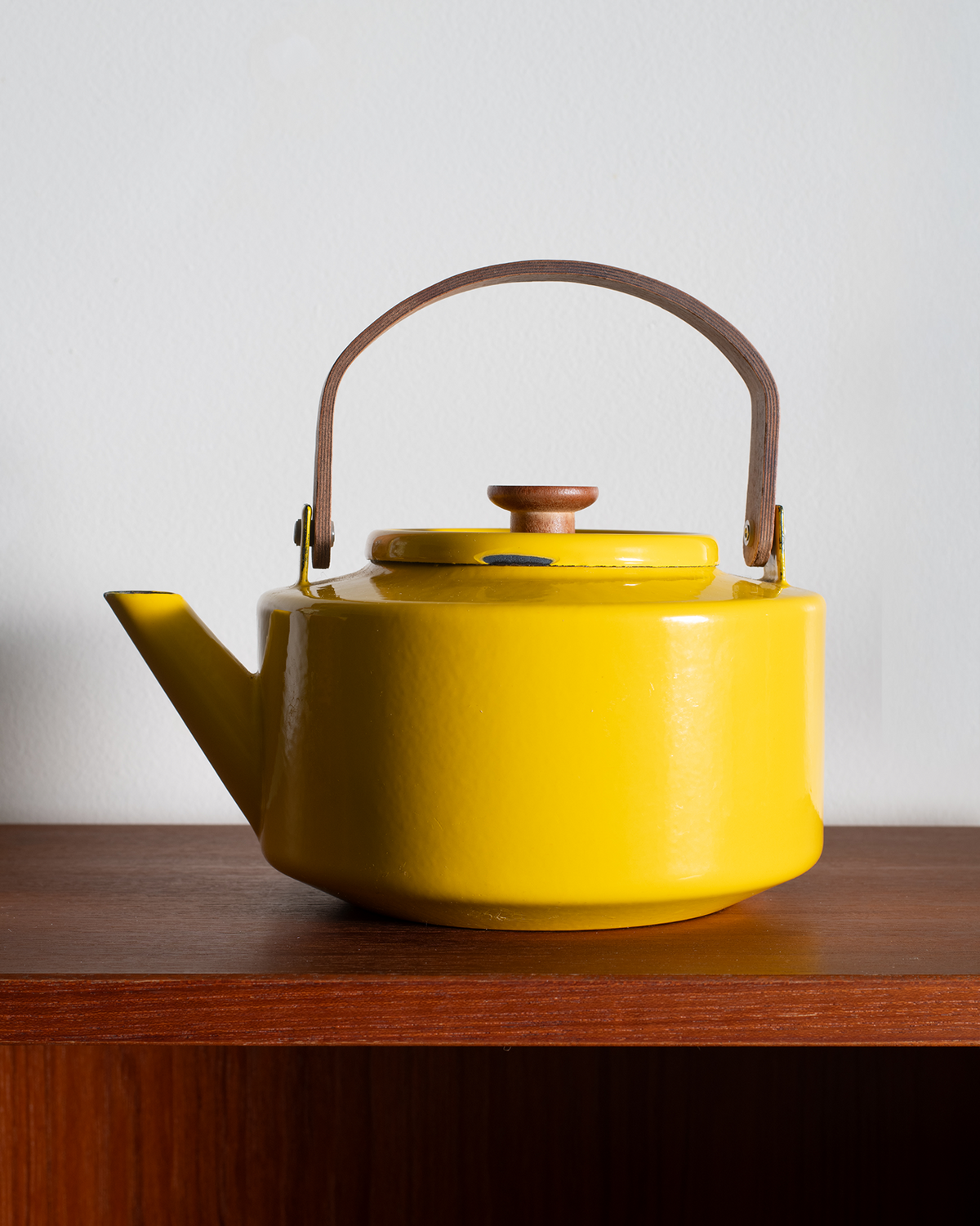 A bright yellow enamel teapot with a brown leather handle and wooden lid knob, sitting on a dark wooden shelf against a plain light gray wall.