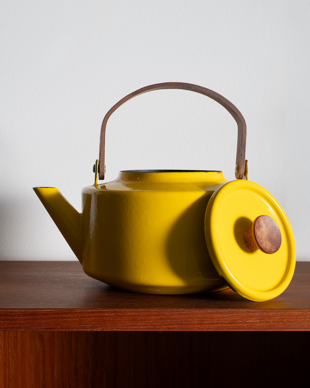 A bright yellow enamel kettle with a brown leather handle and spout trim, shown with its matching lid leaning against it on a wooden shelf against a light gray wall.