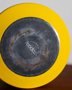 Close-up of the bottom of a bright yellow ceramic plate or dish, showing a dark glazed center with visible wear and a white printed logo reading 'cococo' in the middle.