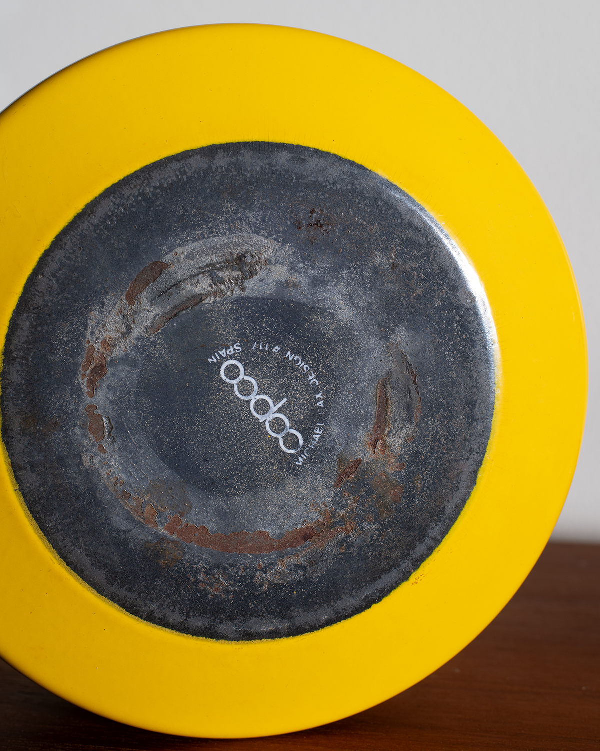 Close-up of the bottom of a bright yellow ceramic plate or dish, showing a dark glazed center with visible wear and a white printed logo reading 'cococo' in the middle.