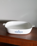 A white ceramic baking dish with side handles and a blue floral pattern, resting on a dark wooden surface against a plain light gray wall.
